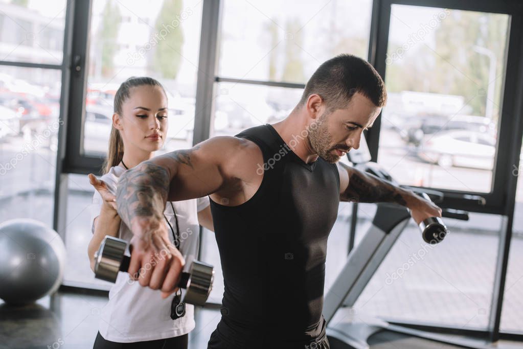 female personal trainer helping sportsman to do exercises with dumbbells at gym