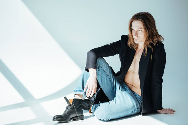 handsome shirtless man with long hair posing in black jacket and jeans, on grey