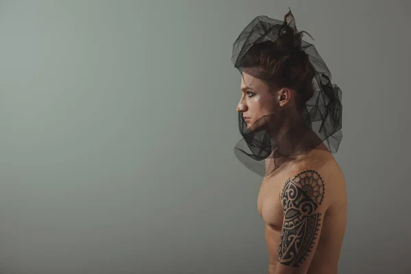 shirtless tattooed man posing with black net on head for fashion shoot, isolated on grey