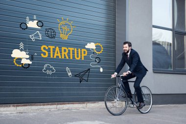 smiling young businessman in suit riding bicycle on street with startup inscription and business icons on gate entrance clipart