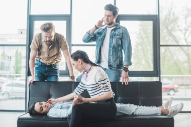 Business colleagues helping young fainted woman lying on sofa in office