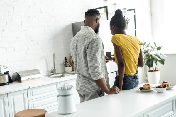 キッチンでスマート フォンで探しているアフリカ系アメリカ人のカップルの背面図 — ストック写真