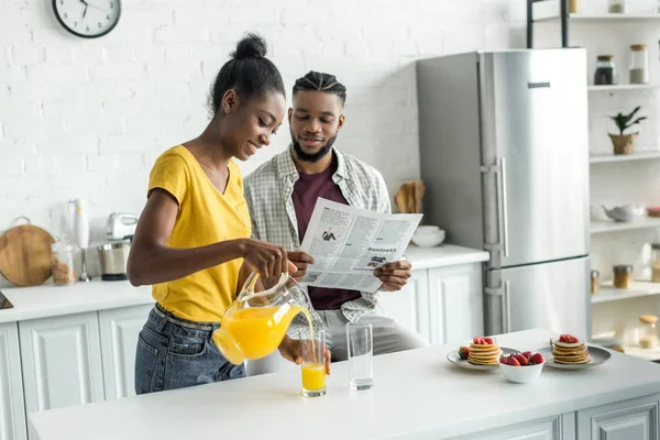 アフリカ系アメリカ人の彼氏が読む新聞とガール フレンドの台所でガラスのオレンジ ジュースを注ぐ — ストック写真