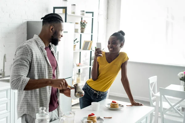 キッチンでスマート フォンでのボーイ フレンドの写真を撮って笑顔のアフリカ系アメリカ人のガール フレンド  — 無料ストックフォト