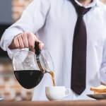 Abgeschnittenes Bild eines Einzelgängers, der Kaffee einschenkt und sein Smartphone in der Küche hält