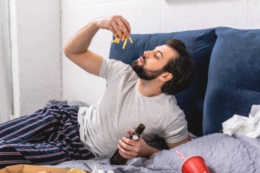 handsome loner eating french fries and holding bottle of beer in bedroom clipart