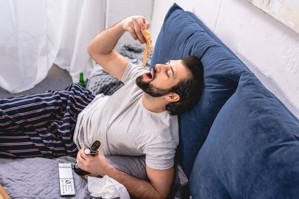 high angle view of handsome loner eating pizza and holding bottle of beer in bedroom
