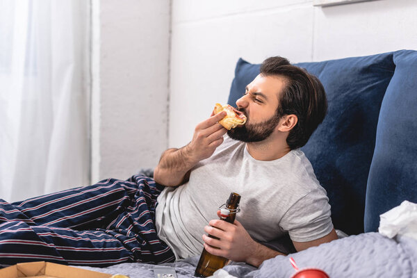 handsome loner eating pizza and holding bottle of beer in bedroom