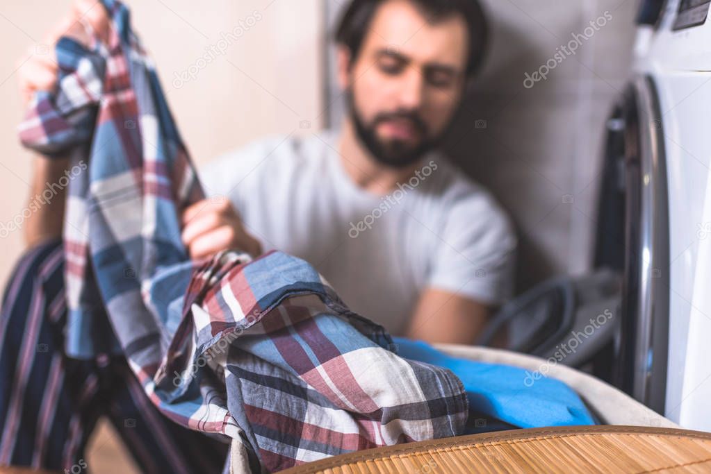 handsome loner taking clothes for washing near washing machine in bathroom with shirt on foreground