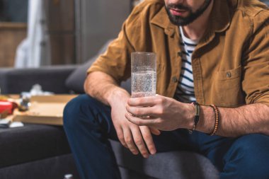 cropped image of loner with hangover holding glass of water at living room clipart