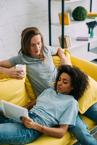 Junges Multiethnisches Paar Auf Sofa Mit Kaffeetasse Und Digitalem Tablet — kostenloses Stockfoto