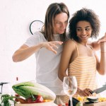 Smiling african american woman holding boyfriend while he gesturing by hands at kitchen