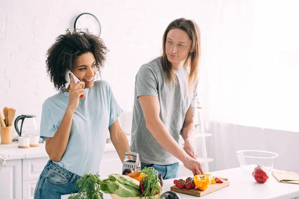 男人切蔬菜 看着微笑的非洲裔美国女友谈论智能手机在厨房 — 图库照片