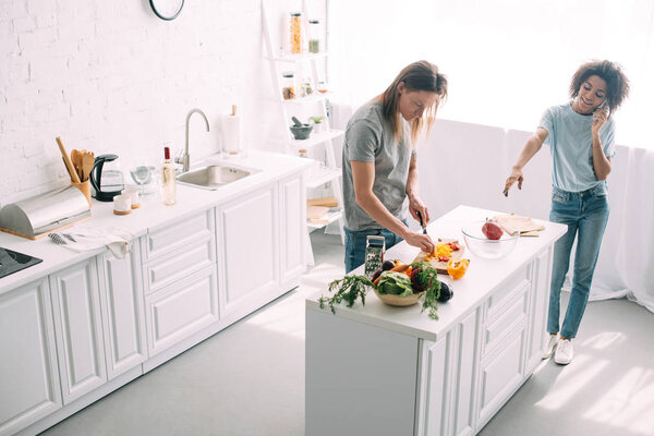 high angle view of young woman talking on smartphone and pointing by hand to boyfriend while he cutting vegetables at kitchen 