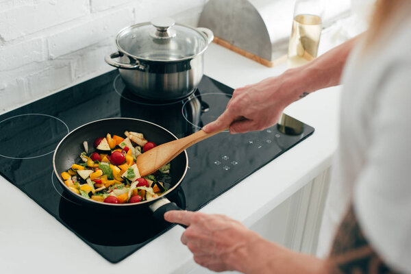 обрезанный снимок человека, готовящего овощи на сковородке на электрической плите
