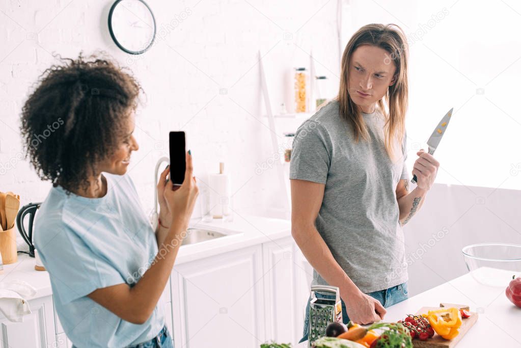 smiling african american woman shooting boyfriend posing with knife on smartphone