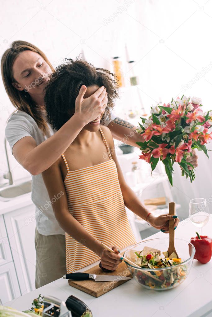 man covering eyes of girlfriend from behind and preparing to give her flowers at kitchen 