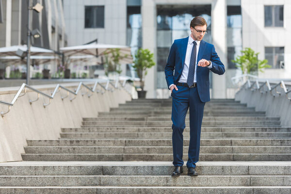 handsome young businessman in stylish suit standing on stairs near business building and looking at watch
