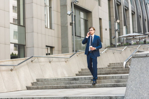 businessman with coffee to go talking by phone while walking by stairs near business building