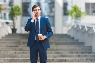 gülümseyen genç işadamı kahve ile şık elbiseli telefonla iş Binası yakınındaki merdivenlerde konuşmaya başladın