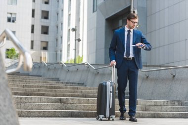 handsome young businessman with luggage and flight tickets walking by business district and looking at watch clipart