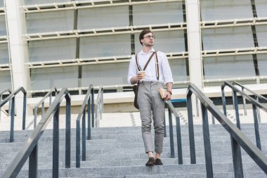 attractive young man walking down stairs in front of stadium with book and coffee to go clipart