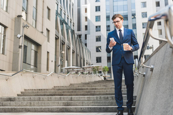 handsome young businessman in stylish suit with paper cup of coffee using smartphone near business building