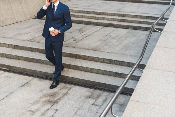 cropped shot of businessman in stylish suit with coffee to go talking by phone while going down stairs