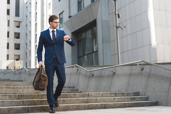attractive young businessman in stylish suit with briefcase looking at watch in business district