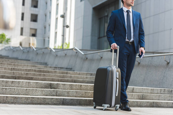 cropped shot of businessman with luggage and flight tickets walking by business district