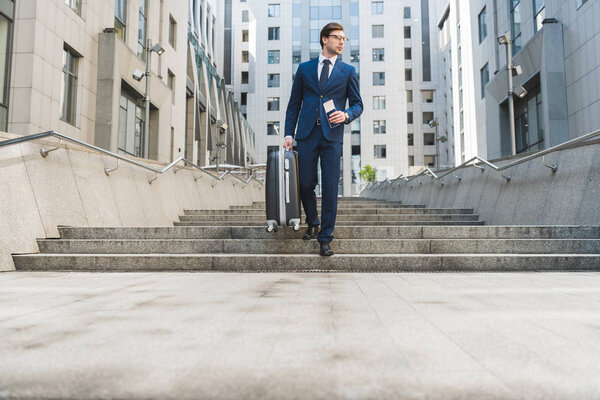 привлекательный молодой бизнесмен в стильном костюме с багажом и авиабилетами спускающийся по лестнице в деловой район
