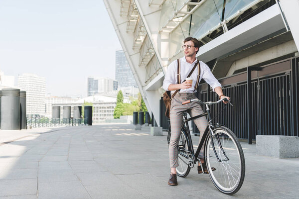 handsome young man with paper cup of coffee on vintage bicycle near sports stadium building