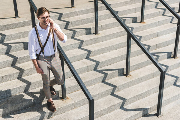 high angle view of handsome young man with folded laptop walking on stairs on city street and talking by phone
