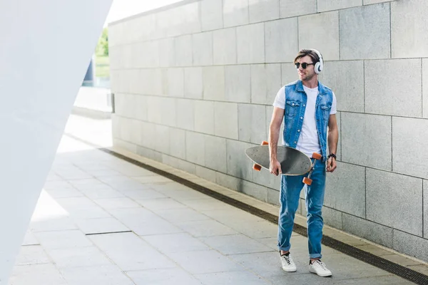 Улыбающийся Молодой Человек Наушниках Скейтбордом Идущий Улице — стоковое фото