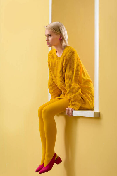 вид сбоку стильной женщины в розовых туфлях, желтом свитере и колготках, сидящей на декоративном витрине
