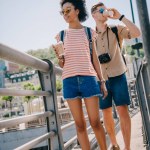 Межрасовая пара туристов с одноразовыми чашками кофе и камерой ходьба по мосту