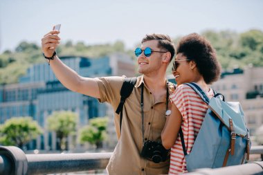 multiethnic couple of travelers in sunglasses taking selfie on smartphone 