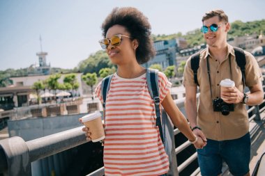turistler ırklararası çift kağıt bardak kahve ve elele ve köprü üzerinde yürüme kamera ile