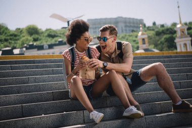 young male traveler trying to eat girlfriend croissant on stairs  clipart