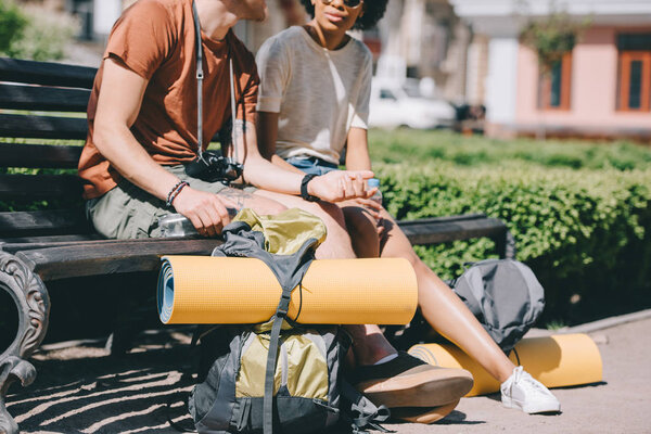 cropped image of couple of travelers sitting on bench with backpacks and mats 