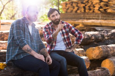 bearded lumberjack pointing by finger to sitting colleague at sawmill  clipart