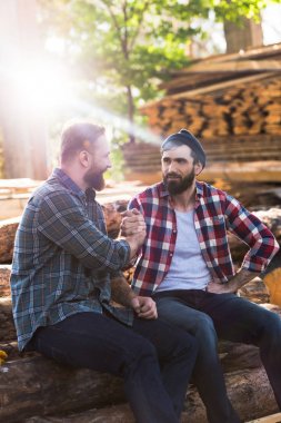 bearded lumberjacks shaking hands of each other and sitting on logs at sawmill  clipart