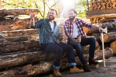 smiling lumberjack with coffee cup pointing by finger to partner with axe sitting on logs at sawmill  clipart