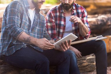 cropped image of bearded lumberjacks sitting on logs with axe and digital tablet at sawmill  clipart