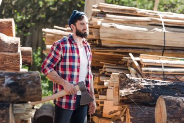 bearded lumberjack in checkered shirt standing with axe at sawmill  clipart