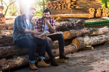 two lumberjacks sitting on logs with axe and using digital tablet at sawmill  clipart