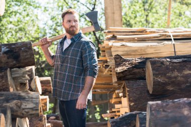 lumberjack in checkered shirt holding axe on shoulder between logs at sawmill   clipart