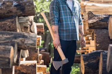 cropped image of lumberjack in checkered shirt with tattooed hands holding axe between logs at sawmill  clipart