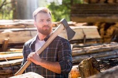lumberjack in checkered shirt holding axe on shoulder at sawmill clipart