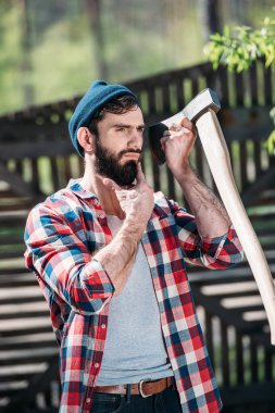 lumberjack in checkered shirt shaving beard by axe at sawmill  clipart